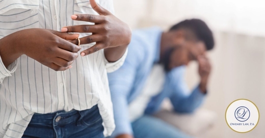 Woman taking off her wedding band and man in the backgound sitting on a couch with his hand on his head.